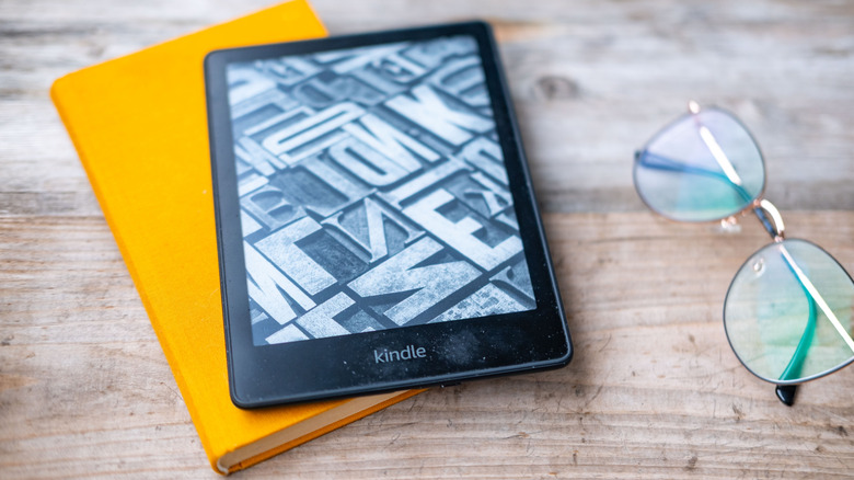 A Kindle e-reader next to a pair of glasses and a physical book