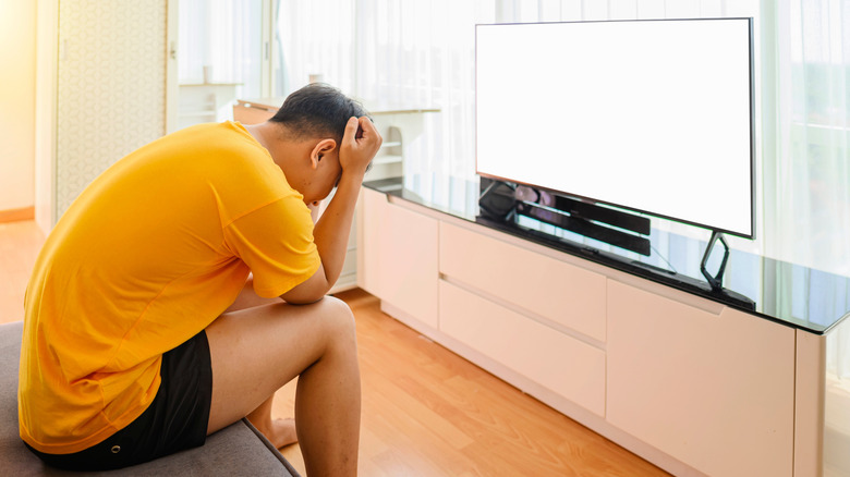 A man with a yellow shirt with hands on head near a tv with a blank white picture