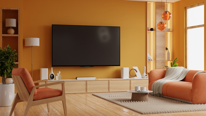 A tv hanging on an orange wall with dark orange couches in the foreground