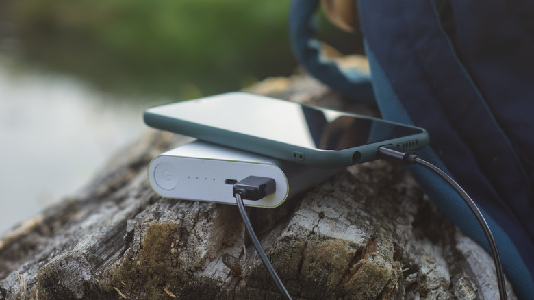 A smartphone plugged into a power bank that's placed on a log and next to a blue backpack