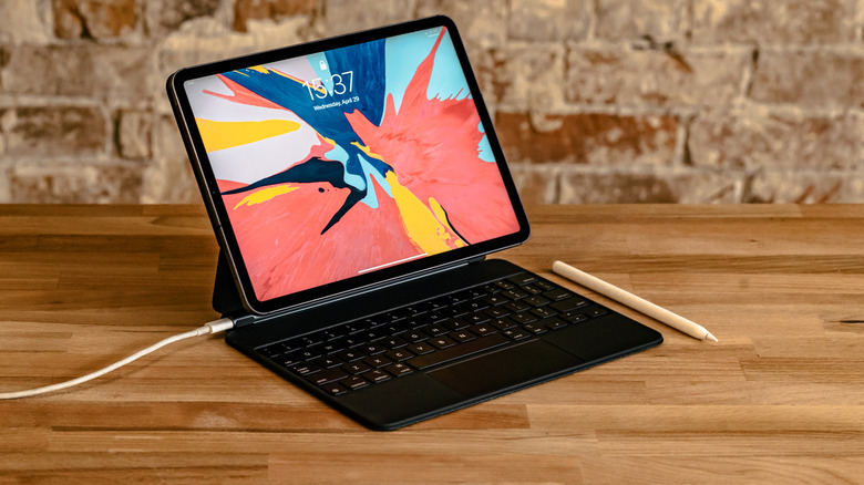 Apple iPad attached to the Magic Keyboard and charging with a white USB-C cord/cable on a wooden desk with an Apple Pencil on the side.