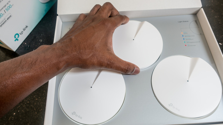 A hand reaching to grab a round mesh Wi-Fi router from a box of three nodes