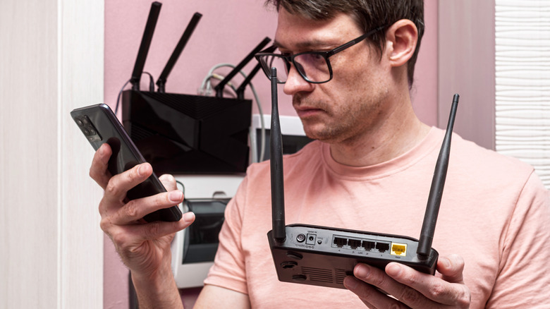A man holding a router in one hand while looking at a phone he is holding in the other hand