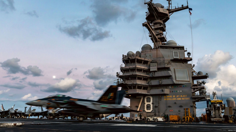 An F/A-18F Super Hornet lands on the Gerald R Ford at sunset, its mast featuring prominently against the sky.