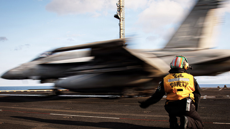 A crouching sailor in yellow safety vest signals a speeding F/A-18E Super Hornet to launch from the USS Gerald R Ford via its electromagnetic catapult