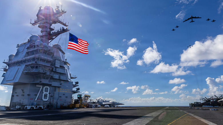 A fleet of jets soar over the U.S.S Ford on a sunny day, as the American flag waves above several docked jets and helicopters on its deck.