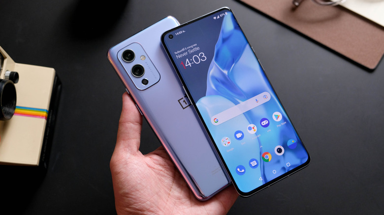 a person holding a OnePlus 9 showing the "Never Settle" slogan of OxygenOS on the screen