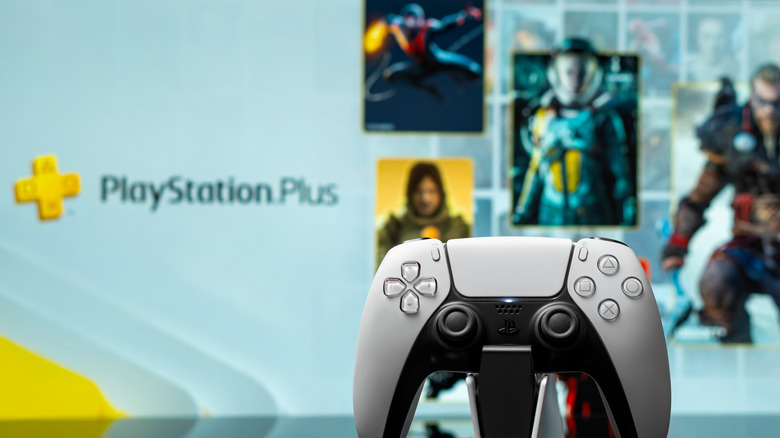 A picture of a DualSense controller on a charging dock, with the PS Plus hero image in the background showing various games available on the service