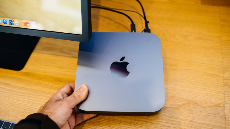A hand grasps the corner of an Apple Mac Mini computer atop a tan brown desk.