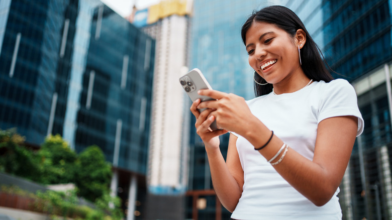 A young woman outside, smiling while she looks at her phone.