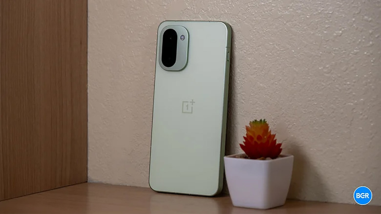 A OnePlus 15R phone leaning against a wall beside a plant.