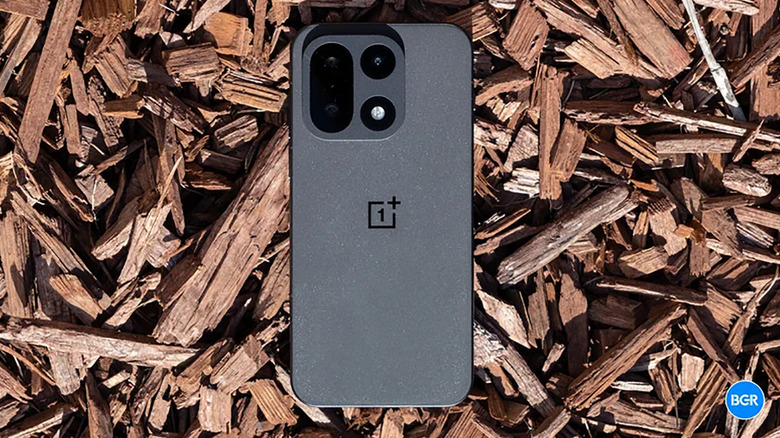 A OnePlus 15 phone face down on some wood chips.