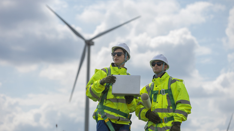 Two people in yellow jackets and hard hats inspecting a wind farm with a turbine behind them