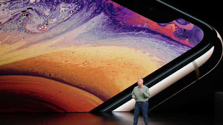 Apple's Phil Schiller on stage during the introduction of the iPhone XS series in 2018, with a stainless steel iPhone XS on a screen in the background.