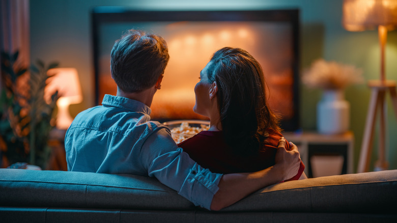 Couple sitting close together on a couch watching a movie on a softly lit living-room television