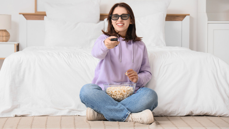 Woman wearing 3D glasses sitting cross-legged with popcorn and pointing a remote toward the TV