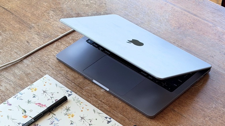 Half-opened MacBook Pro on a table with a little notebook and a pen underneath it.