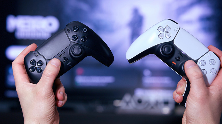 A person holding a black PS5 DualSense controller in one hand and a white one in the other, with a TV blurred in the distance