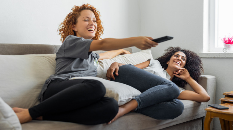 Two young women on a couch watching TV with a remote in hand