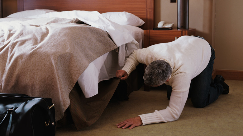 A man looks under a bed in a hotel room.