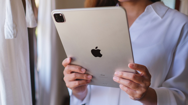 An individual in a white button-up shirt holds a silver iPad between both their hands, the image is cropped so that the user's face cannot be seen and there is an emphasis on the iPad