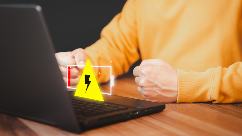 An individual in a loud, orange sweater sits behind a black laptop, a computer-generated icon showing a battery with a warning sign floats above the black laptop