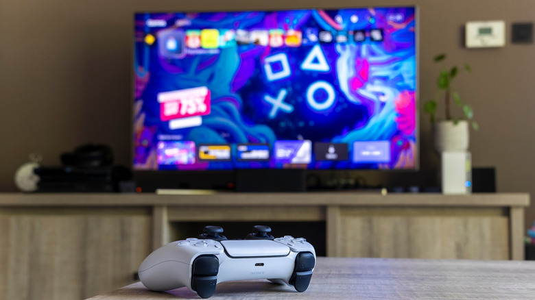 A white PlayStation 5 controller sits on a coffee table, in the background is a television displaying the PS5 Home screen