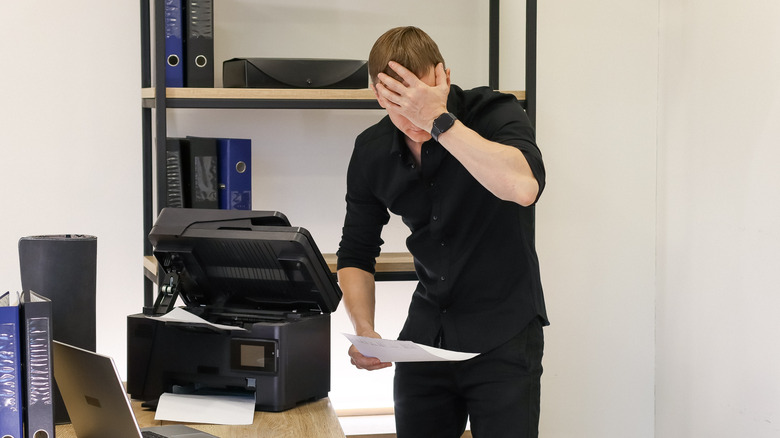 A person holding a sheet of paper, frustrated with a broken black printer in an office environment