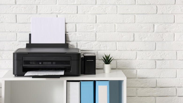 A black printer with sheets loaded in the paper tray placed on a white table against a white brick wall in an office