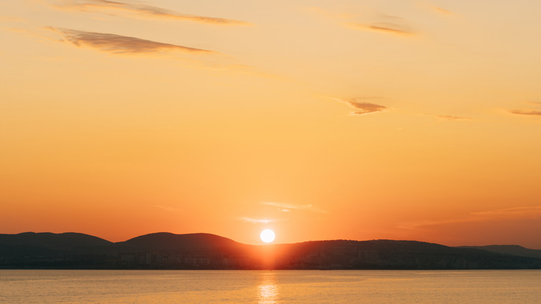 The sun setting over a lake and hills, with some faint clouds in the orange sky