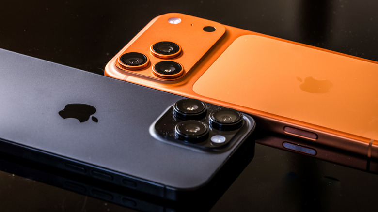 A black iPhone 17 lying alongside an orange iPhone 17 Pro, both of which have prominent camera bumps
