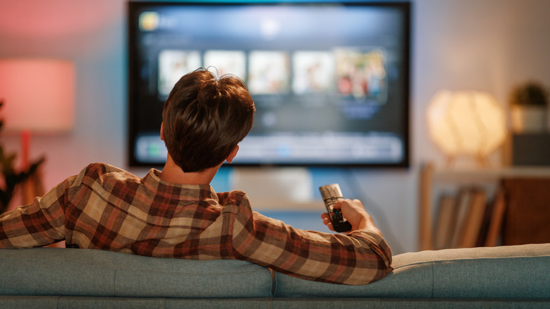 The back of a man on a couch holding a remote in front of a TV.
