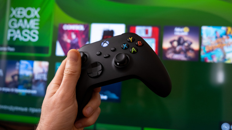 A hand holding an Xbox Controller with the Xbox home screen in the background