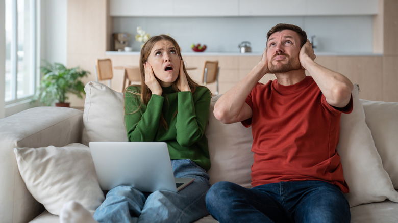 Frustrated couple sitting on a couch with a laptop, covering their ears as they look upward in annoyance.