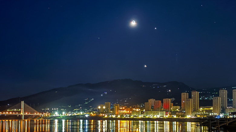 The crescent moon is seen with the planets Jupiter (lower right) and Venus (bottom), above the Yangtze River and the illuminated city skyline on August 20, 2025 in Chongqing, China.
