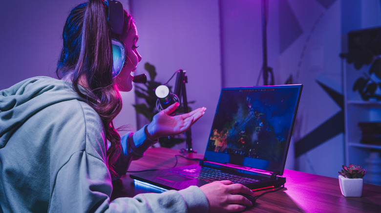 A woman gaming on her laptop, having a multiplayer game open on the screen and talking into a headphone mic