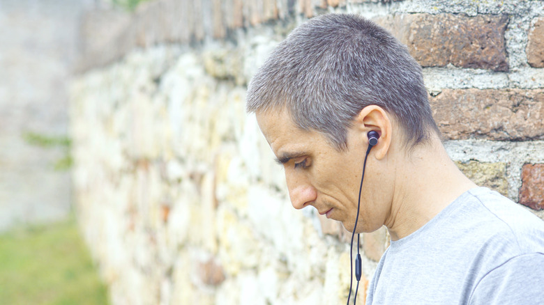 A man standing against a wall wearing wired earbuds.