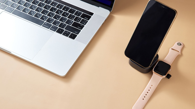 A phone and smartwatch on a wireless charger beside a laptop.