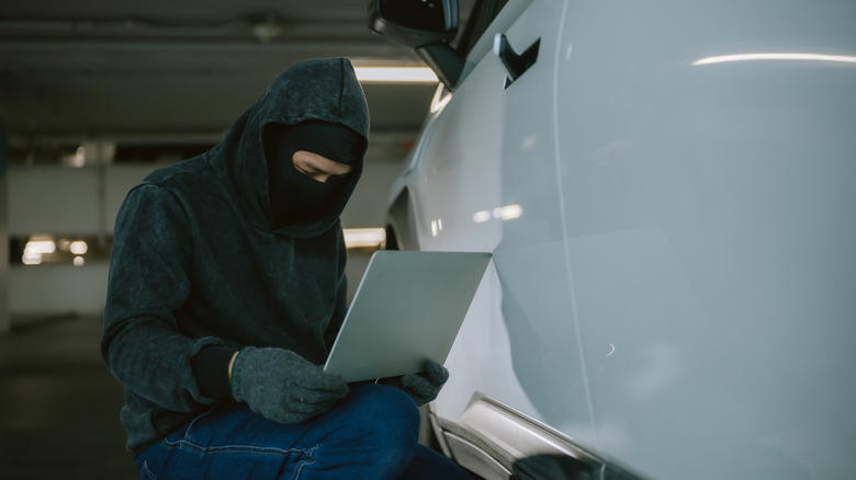 A person dressed in dark clothing and gloves holding a laptop is crouched near a parked vehicle