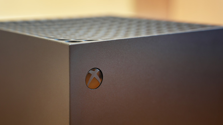A closeup of the "X" on an Xbox Series X as the device sits in a room that is too blurred by camera effects to be made out completely