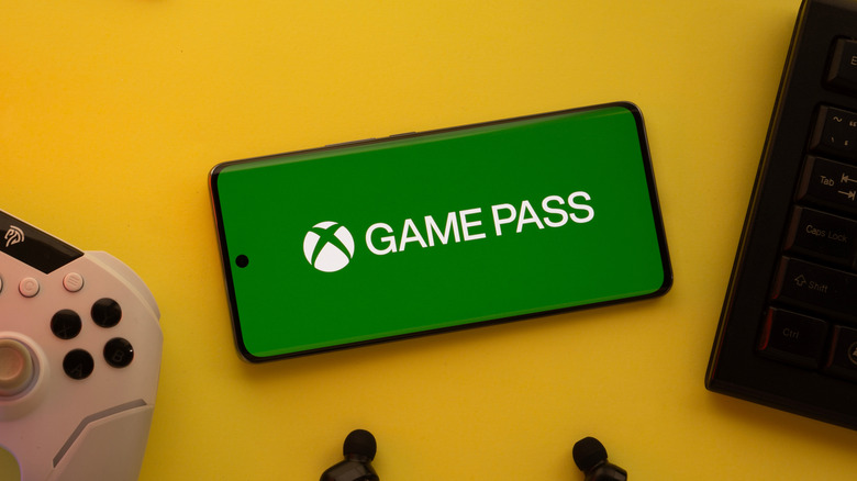 A cellphone displays the Xbox Game Pass logo as it sits on a yellow backdrop between a gaming controller and another device