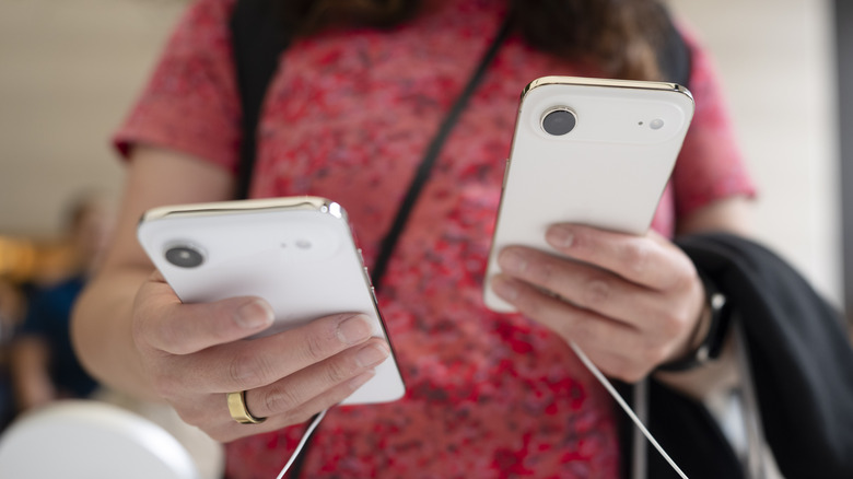 A woman in a red floral shirt holding two different white iPhone 17 Airs inside of an Apple Store, security cables dangle from the phones