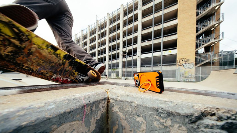 A Yeti LoadOut iPhone case in orange propped up by its kickstand, a skateboarder beside it.