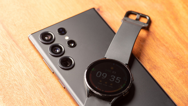A Samsung Galaxy Watch with a black wrist band sits on top of a black Samsung Galaxy smartphone, both are on top of a wooden surface