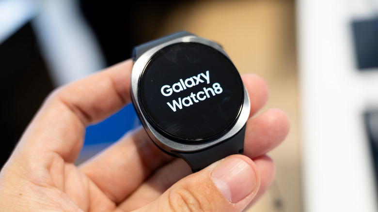 A Samsung Galaxy Watch 8 with a black band rests over the fingers of a user's left hand, the background is blurred heavily but it vaguely resembles an electronics store