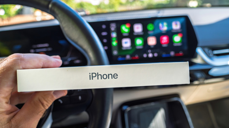 A person sitting in a car and holding a boxed iPhone in front of a CarPlay infotainment screen.