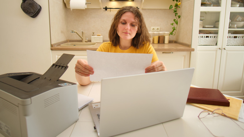 Woman inspecting a sheet of paper with an open laptop and desktop printer nearby.