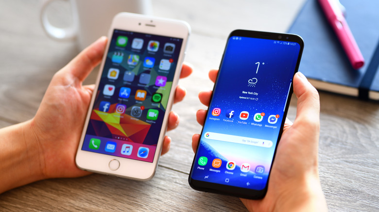 A pair of hands holding an iPhone and an Android device on a light office. Both devices show the home screen.