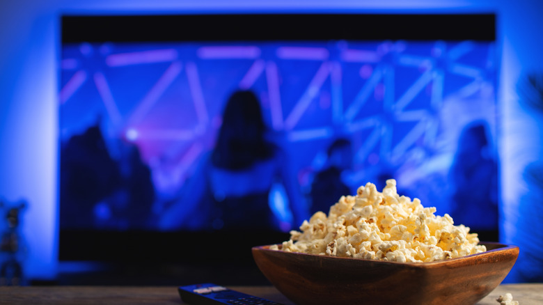 A wooden bowl of popcorn placed on a table in front of a TV screen.