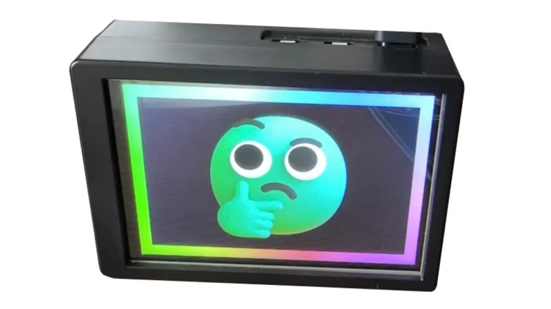 Un pequeño dispositivo de escritorio con pantalla integrada muestra un emoji pensativo sobre un fondo iluminado con bordes de colores. La carcasa del dispositivo es negra y compacta.